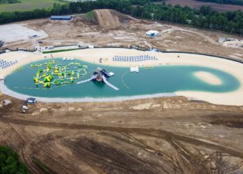 A high-angle aerial view capturing the expansive SkyLake Adventures water and adventure park during its final construction and build-out phases in Sheridan, Indiana. At the center lies a large, kidney-shaped man-made lake filled with vibrant, turquoise-colored water, completely lined by a wide border of light sand beaches. Floating on the left side of the lake is a massive, sprawling bright yellow and green inflatable obstacle course, with a separate multi-tiered gray and blue water slide structure positioned right next to it. The immediate surrounding landscape consists of heavily graded brown dirt, tire tracks, and undergoing earthworks, with a large tubing hill mound rising in the far background. Rows of blue lounge chairs and white umbrellas stand neatly organized along the upper shoreline.