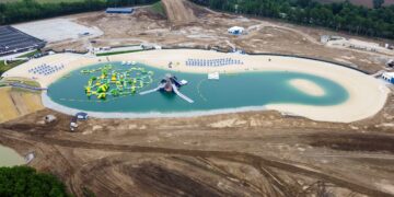 A high-angle aerial view capturing the expansive SkyLake Adventures water and adventure park during its final construction and build-out phases in Sheridan, Indiana. At the center lies a large, kidney-shaped man-made lake filled with vibrant, turquoise-colored water, completely lined by a wide border of light sand beaches. Floating on the left side of the lake is a massive, sprawling bright yellow and green inflatable obstacle course, with a separate multi-tiered gray and blue water slide structure positioned right next to it. The immediate surrounding landscape consists of heavily graded brown dirt, tire tracks, and undergoing earthworks, with a large tubing hill mound rising in the far background. Rows of blue lounge chairs and white umbrellas stand neatly organized along the upper shoreline.