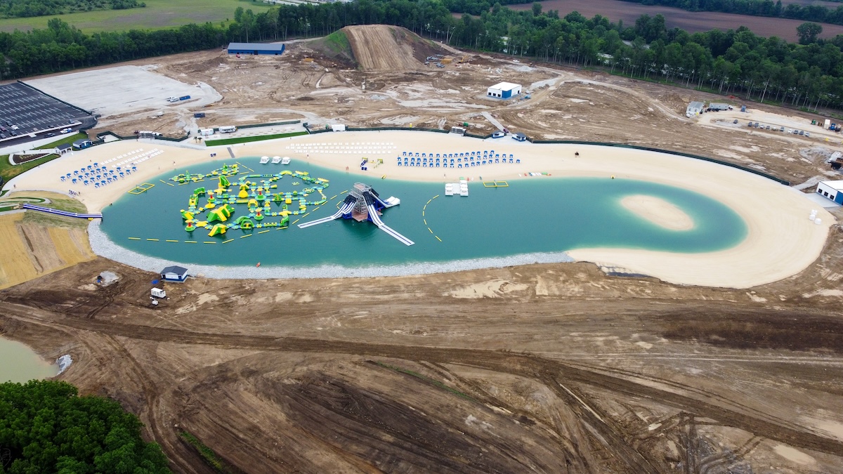 A high-angle aerial view capturing the expansive SkyLake Adventures water and adventure park during its final construction and build-out phases in Sheridan, Indiana. At the center lies a large, kidney-shaped man-made lake filled with vibrant, turquoise-colored water, completely lined by a wide border of light sand beaches. Floating on the left side of the lake is a massive, sprawling bright yellow and green inflatable obstacle course, with a separate multi-tiered gray and blue water slide structure positioned right next to it. The immediate surrounding landscape consists of heavily graded brown dirt, tire tracks, and undergoing earthworks, with a large tubing hill mound rising in the far background. Rows of blue lounge chairs and white umbrellas stand neatly organized along the upper shoreline.