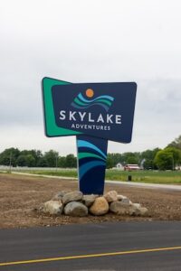 A vertical, eye-level shot capturing the official entrance sign for SkyLake Adventures in Sheridan, Indiana. The modern pylon sign is positioned next to an asphalt road with a yellow line. The top portion of the sign features a dark blue, asymmetrical geometric panel layered over a bright green background panel. On the dark blue face, the words "SKYLAKE ADVENTURES" are written in bold, white sans-serif capital lettering beneath a logo depicting wavy teal lines and an orange sun. The vertical support pillar is decorated with curving blue, teal, and navy stripes. The entire base of the sign is surrounded by an arrangement of large, rounded natural river stones and boulders on a patch of brown soil, with green fields, trees, and a distant red barn under a cloudy sky.