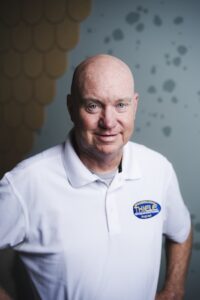 A portrait of a bald, smiling man wearing a white polo shirt with a blue and yellow Thiele Heating & Air Conditioning logo on the left chest, posing against a gray textured wall.
