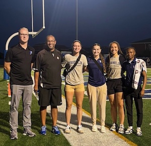 Night view on a brightly lit football field turf. Six people pose shoulder-to-shoulder under a white-and-yellow goalpost. From left to right: a tall man in a black polo shirt and grey trousers; a man wearing a black short-sleeve polo shirt and black athletic shorts; a woman in a grey Franklin College shirt and yellow shorts; a woman in a navy blue polo shirt and khaki slacks; a woman in a grey Franklin College t-shirt and black athletic shorts; and a woman wearing a navy blue polo shirt and dark pants with a white towel draped over her shoulder. The dark night sky and the outline of stadium bleachers are visible in the background.