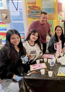 Indoor shot of a group participating in an art or craft workshop. In the foreground, two young women sit side-by-side smiling at the camera. The woman on the left wears blue protective gloves and holds a clear plastic cup. The woman beside her wears a white long-sleeve shirt with the text "Hailey's ARMY" printed across the front. Behind them, a man in a maroon t-shirt leans forward with a smile. To the right, another woman sits next to a large pink insulated travel mug. The table is draped in a black plastic sheet and scattered with small white paper cups, water bottles, and small wooden canvas boards showing colorful paint designs. The colorful background features rustic wood signs with painted words.