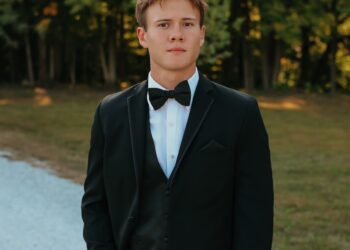A medium, three-quarter portrait of a young man with short brown hair posing outdoors. He is dressed in a formal black tuxedo suit with a matching vest, a crisp white button-down shirt, and a classic black bow tie. He stands with one hand slipped casually into his pants pocket, looking directly forward with a calm expression. The background features a white gravel path next to a grassy area and a soft-focus line of green trees lit by warm, late-afternoon sunlight.