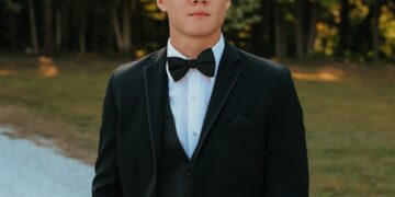 A medium, three-quarter portrait of a young man with short brown hair posing outdoors. He is dressed in a formal black tuxedo suit with a matching vest, a crisp white button-down shirt, and a classic black bow tie. He stands with one hand slipped casually into his pants pocket, looking directly forward with a calm expression. The background features a white gravel path next to a grassy area and a soft-focus line of green trees lit by warm, late-afternoon sunlight.