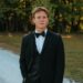 A medium, three-quarter portrait of a young man with short brown hair posing outdoors. He is dressed in a formal black tuxedo suit with a matching vest, a crisp white button-down shirt, and a classic black bow tie. He stands with one hand slipped casually into his pants pocket, looking directly forward with a calm expression. The background features a white gravel path next to a grassy area and a soft-focus line of green trees lit by warm, late-afternoon sunlight.