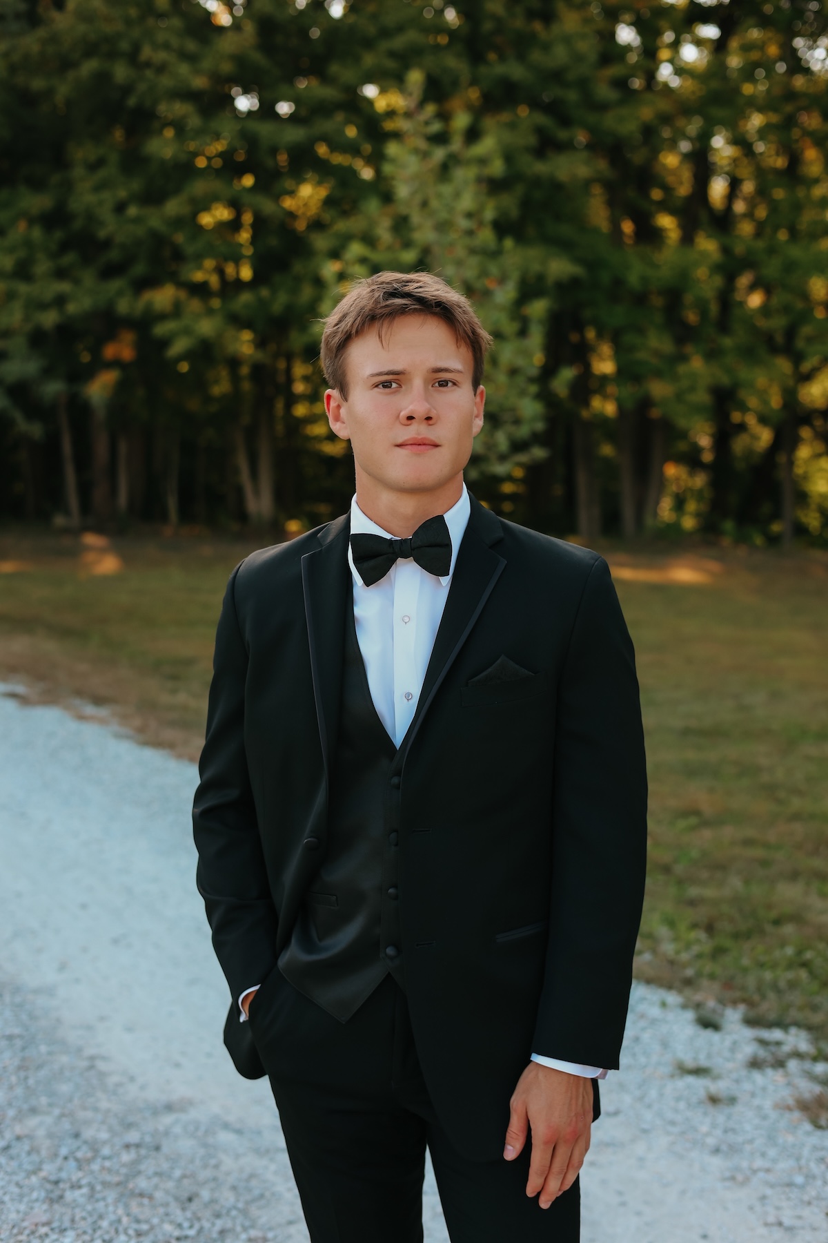 A medium, three-quarter portrait of a young man with short brown hair posing outdoors. He is dressed in a formal black tuxedo suit with a matching vest, a crisp white button-down shirt, and a classic black bow tie. He stands with one hand slipped casually into his pants pocket, looking directly forward with a calm expression. The background features a white gravel path next to a grassy area and a soft-focus line of green trees lit by warm, late-afternoon sunlight.