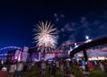A night photograph captured from a low angle shows a large crowd gathered on a grassy field watching a vibrant fireworks display burst over a large steel bridge. The bridge is illuminated with red lights on the right section, while a neighboring bridge to the left is lit with blue. An outdoor stage with an American flag graphic stands on the left side of the field, and bright event tents line the background under a dark, partly cloudy night sky.