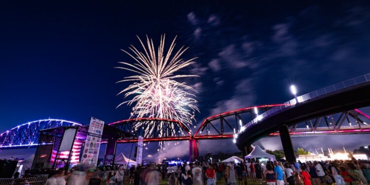 A night photograph captured from a low angle shows a large crowd gathered on a grassy field watching a vibrant fireworks display burst over a large steel bridge. The bridge is illuminated with red lights on the right section, while a neighboring bridge to the left is lit with blue. An outdoor stage with an American flag graphic stands on the left side of the field, and bright event tents line the background under a dark, partly cloudy night sky.