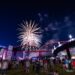 A night photograph captured from a low angle shows a large crowd gathered on a grassy field watching a vibrant fireworks display burst over a large steel bridge. The bridge is illuminated with red lights on the right section, while a neighboring bridge to the left is lit with blue. An outdoor stage with an American flag graphic stands on the left side of the field, and bright event tents line the background under a dark, partly cloudy night sky.