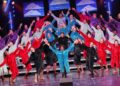 A large show choir performs on a multi-tiered stage, with dozens of singers dressed in oversized, plush hooded robes in a variety of solid colors including red, purple, light blue, teal, gray, and black. The performers stand barefoot with their arms outstretched in a dramatic, expressive pose. Three video monitors hanging in the background display stylized images of white clouds against a blue sky, while theater spotlights illuminate the stage.