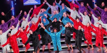 A large show choir performs on a multi-tiered stage, with dozens of singers dressed in oversized, plush hooded robes in a variety of solid colors including red, purple, light blue, teal, gray, and black. The performers stand barefoot with their arms outstretched in a dramatic, expressive pose. Three video monitors hanging in the background display stylized images of white clouds against a blue sky, while theater spotlights illuminate the stage.