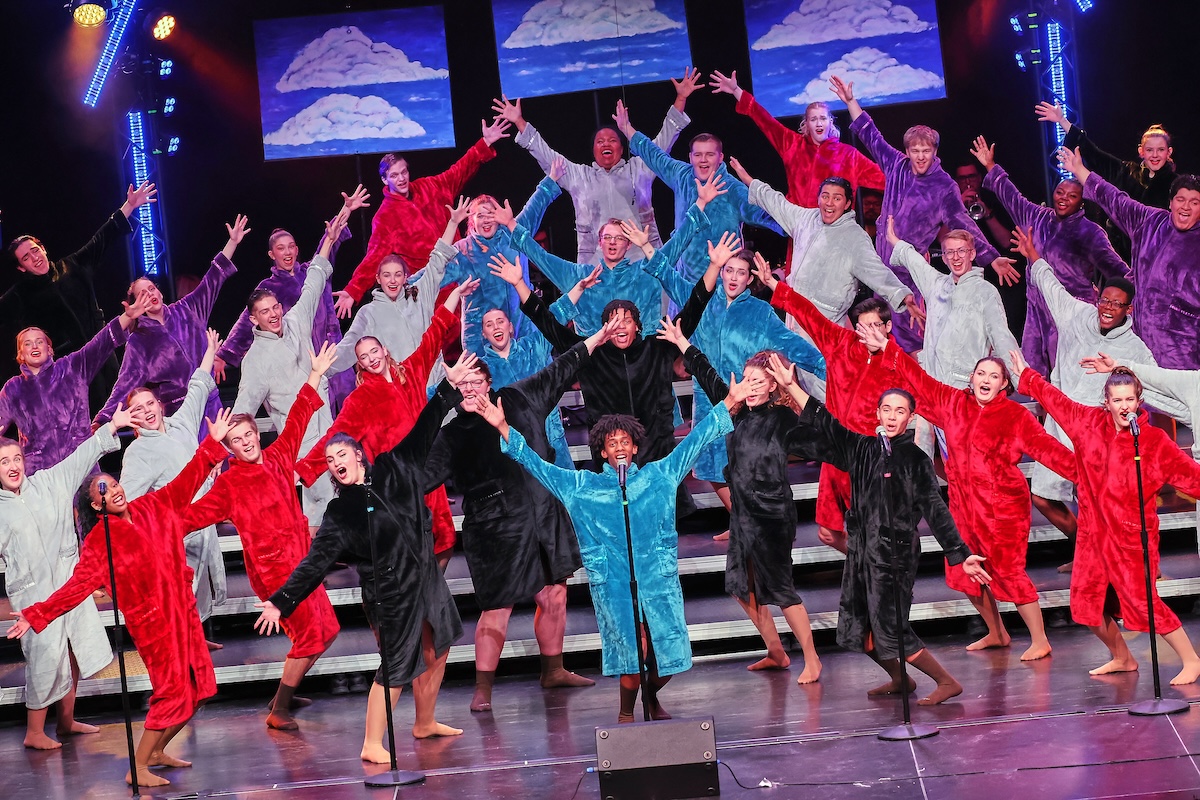 A large show choir performs on a multi-tiered stage, with dozens of singers dressed in oversized, plush hooded robes in a variety of solid colors including red, purple, light blue, teal, gray, and black. The performers stand barefoot with their arms outstretched in a dramatic, expressive pose. Three video monitors hanging in the background display stylized images of white clouds against a blue sky, while theater spotlights illuminate the stage.