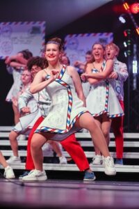 A female show choir performer with dark hair and red lipstick smiles brightly while hitting a dynamic dance pose on a tiered stage. She wears a white sleeveless A-line dress featuring red and blue diagonal candy-stripe borders resembling vintage airmail envelopes, paired with white sneakers. Several other student performers dance on the risers behind her under theater stage lighting, with stylized mail-themed props visible in the background.