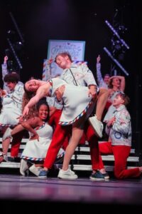 A male show choir performer in a white patterned jacket and red pants dips a female performer on a tiered stage. The female dancer wears a white sleeveless dress with red and blue diagonal stripe trim and white sneakers, smiling mid-dip. In the background, other members of the ensemble sing and react dynamically on the stage risers under overhead theater lighting next to a sign that reads "Just Married".