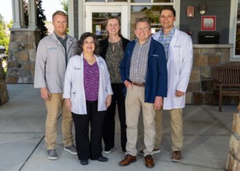 Five medical professionals pose together outside the entrance of Fall Creek Veterinary Medical Center. Two women and three men stand in a group on a concrete walkway in front of glass entry doors. The staff members are dressed in business casual attire, medical lab coats, and clinic jackets embroidered with the company logo. An AAHA Accredited sign is visible on the stone and siding exterior wall to the right.