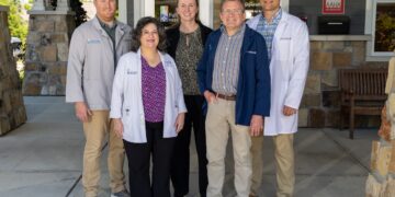 Five medical professionals pose together outside the entrance of Fall Creek Veterinary Medical Center. Two women and three men stand in a group on a concrete walkway in front of glass entry doors. The staff members are dressed in business casual attire, medical lab coats, and clinic jackets embroidered with the company logo. An AAHA Accredited sign is visible on the stone and siding exterior wall to the right.