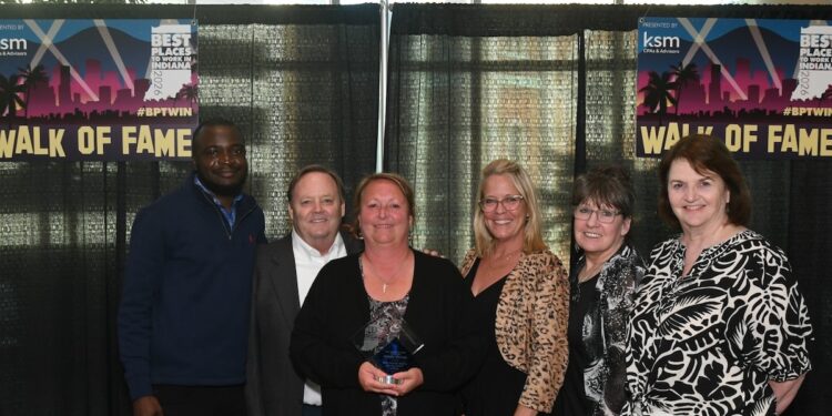 A group of six colleagues stand smiling together in front of a black curtain backdrop at an awards event. A woman in the center holds a glass trophy award. Behind them, large banners read "Best Places to Work in Indiana 2026", "Presented by KSM CPAs & Advisors", and "Walk of Fame" with the hashtag #BPTWIN.