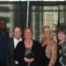 A group of six colleagues stand smiling together in front of a black curtain backdrop at an awards event. A woman in the center holds a glass trophy award. Behind them, large banners read "Best Places to Work in Indiana 2026", "Presented by KSM CPAs & Advisors", and "Walk of Fame" with the hashtag #BPTWIN.