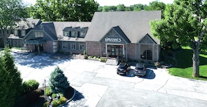A high-angle, wide aerial drone photograph capturing the exterior of the new Brianne's Boutique storefront location in Greenwood, Indiana. The building features a large, single-story brick structure with multiple steep, brown-shingled gabled roofs and dark dormer windows. Positioned directly over the central glass entry doors is a prominent white sign reading "BRIANNE'S". In front of the shop, a vast, light gray concrete parking lot spans the foreground, with two dark-colored SUVs parked near the main entrance steps. Mature green trees and lush landscaping surround the property under bright, direct sunlight.