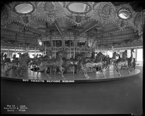 A archival black-and-white photograph showcases the 1922 William Dentzel Deluxe Menagerie Carousel platform wdrb.com. The grand, ornate merry-go-round features rows of elaborately hand-carved wooden animals, including jumping horses and a tall giraffe on the outer ring. The base of the rotating platform bears a painted sign reading "Buy Tickets Before Riding." Above, the intricately sculpted canopy is adorned with decorative trim, rosettes, and oval mirrors.