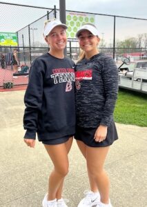 Two teenage female tennis players stand closely together and smile for a photo on an outdoor concrete walkway. Both are wearing white Adidas brand athletic visors and white tennis sneakers. The player on the left has long blonde hair in a ponytail, a dark navy or black crewneck sweatshirt reading "CENTER GROVE TENNIS" with a Trojan logo, and a black tennis skirt. The player on the right has lighter blonde hair and wears a dark gray and black space-dye patterned long-sleeve shirt with a small matching Trojan logo, paired with a black tennis skirt. Behind them, a chain-link fence encloses outdoor courts, with a sign that reads "Spring Tennis Clinics."