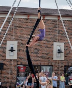 An aerial silk performer hangs upside down in mid-air from long, black fabric panels suspended from a large metal outdoor rig. The female acrobat wears a purple leotard, arching her back with one arm extended while a trainer below holds the base of the fabric to steady it. A crowd of spectators stands in the background watching the exhibition outside a multi-story red brick building with wall-mounted lamps.