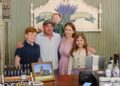 A family of five—a father, mother, two sons, and a daughter—smile together behind a wooden store counter. In the background, a painted sign reads "Bloomfield Lavender Apothecary" with a lavender illustration. The counter displays various small lavender product bottles, a framed picture of a schoolhouse, and a modern point-of-sale checkout system.