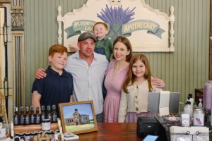 A family of five—a father, mother, two sons, and a daughter—smile together behind a wooden store counter. In the background, a painted sign reads "Bloomfield Lavender Apothecary" with a lavender illustration. The counter displays various small lavender product bottles, a framed picture of a schoolhouse, and a modern point-of-sale checkout system.