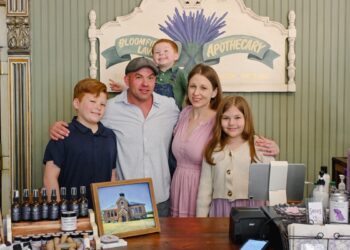 A family of five—a father, mother, two sons, and a daughter—smile together behind a wooden store counter. In the background, a painted sign reads "Bloomfield Lavender Apothecary" with a lavender illustration. The counter displays various small lavender product bottles, a framed picture of a schoolhouse, and a modern point-of-sale checkout system.