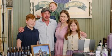 A family of five—a father, mother, two sons, and a daughter—smile together behind a wooden store counter. In the background, a painted sign reads "Bloomfield Lavender Apothecary" with a lavender illustration. The counter displays various small lavender product bottles, a framed picture of a schoolhouse, and a modern point-of-sale checkout system.