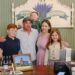 A family of five—a father, mother, two sons, and a daughter—smile together behind a wooden store counter. In the background, a painted sign reads "Bloomfield Lavender Apothecary" with a lavender illustration. The counter displays various small lavender product bottles, a framed picture of a schoolhouse, and a modern point-of-sale checkout system.