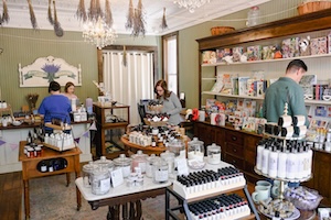 The interior of the Bloomfield Lavender Apothecary shop filled with products. Customers browse central display tables holding jars and bottles, while staff assist people behind a service counter. A large wooden wall cabinet on the right is packed with inventory, and dried lavender bundles hang from the ceiling near a crystal chandelier.