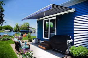 A striped navy blue and white motorized retractable awning extended over a backyard concrete patio. The awning is mounted to a house with blue vinyl siding above a glass sliding door. The patio features wicker chairs with cushions, potted flowers, a covered grill, and a green lawn that stretches toward a lake in the background under a clear blue sky.
