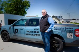 A man stands on a paved driveway next to a light blue Ford Maverick pickup truck with commercial vehicle graphics. The side of the truck displays a "PS PREMIER SHADE" logo and the text "YOUR LOCAL SHADE EXPERT." The truck bed features text reading "Retractable Awnings, Retractable Screens," the phone number (260) 255-6004, and a large QR code.