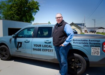 A man stands on a paved driveway next to a light blue Ford Maverick pickup truck with commercial vehicle graphics. The side of the truck displays a "PS PREMIER SHADE" logo and the text "YOUR LOCAL SHADE EXPERT." The truck bed features text reading "Retractable Awnings, Retractable Screens," the phone number (260) 255-6004, and a large QR code.