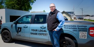 A man stands on a paved driveway next to a light blue Ford Maverick pickup truck with commercial vehicle graphics. The side of the truck displays a "PS PREMIER SHADE" logo and the text "YOUR LOCAL SHADE EXPERT." The truck bed features text reading "Retractable Awnings, Retractable Screens," the phone number (260) 255-6004, and a large QR code.