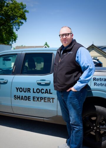 A man stands on a paved driveway next to a light blue Ford Maverick pickup truck with commercial vehicle graphics. The side of the truck displays a "PS PREMIER SHADE" logo and the text "YOUR LOCAL SHADE EXPERT." The truck bed features text reading "Retractable Awnings, Retractable Screens," the phone number (260) 255-6004, and a large QR code.