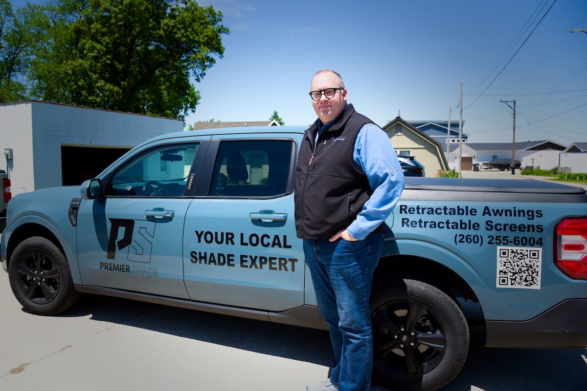 A man stands on a paved driveway next to a light blue Ford Maverick pickup truck with commercial vehicle graphics. The side of the truck displays a "PS PREMIER SHADE" logo and the text "YOUR LOCAL SHADE EXPERT." The truck bed features text reading "Retractable Awnings, Retractable Screens," the phone number (260) 255-6004, and a large QR code.