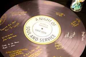 Top-down, slightly angled close-up view of a large prop shaped like a vinyl record, used as a guest book at a formal event. The outer ring is a metallic purple color with textured grooves, entirely covered in handwritten signatures and organization names written in gold paint marker. The white circular center label features a gold border and reads "A NIGHT OF SOUL AND SERVICE" curved along the edge, with "GLITTER & GRATITUDE GALA 2026" printed horizontally in the middle. The corner of a glass jar with colorful paper inside is visible in the upper right background.
