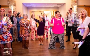 Joyful indoor scene of a crowded dance floor at a community gala. A group of men and women dance under warm lighting, with several people dressed in 1960s or 1970s retro style clothing, including paisley prints, floral dresses, and flared pants. In the foreground, a woman in a bright pink top and patterned black-and-white trousers holds two glowing white foam light sticks. Around her, other guests laugh, dance, and hold matching light sticks. The room features dark wood-look flooring and stacked-stone accent pillars in the background.