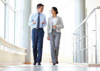 A man and a woman in professional business attire walk side-by-side down a bright, sunlit modern office hallway. The man wears a light blue dress shirt, a striped tie, dark blue trousers, and dress shoes while gesturing with his hands. The woman smiles back at him, wearing a light grey blazer, a white collared shirt, grey dress pants, and black heels.