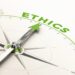 A conceptual close-up illustration of a metallic compass on a white surface. The green arrow of the compass needle points directly toward the word "ETHICS" printed in bold, bright green text along the upper curve of the dial. The word is repeated in fainter gray text along other parts of the ring.