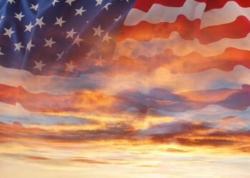 A double-exposure image blending a waving American flag with a dramatic sunset sky filled with glowing orange, yellow, and blue-gray clouds shining through the translucent stars and stripes.