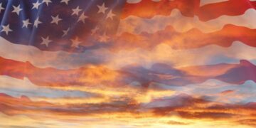 A double-exposure image blending a waving American flag with a dramatic sunset sky filled with glowing orange, yellow, and blue-gray clouds shining through the translucent stars and stripes.