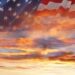 A double-exposure image blending a waving American flag with a dramatic sunset sky filled with glowing orange, yellow, and blue-gray clouds shining through the translucent stars and stripes.