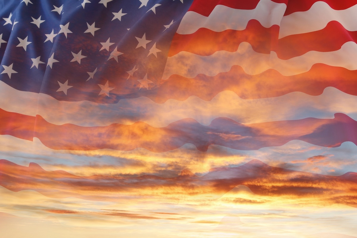 A double-exposure image blending a waving American flag with a dramatic sunset sky filled with glowing orange, yellow, and blue-gray clouds shining through the translucent stars and stripes.