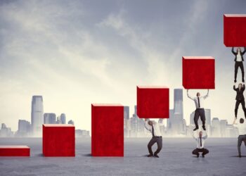 A conceptual digital illustration of professionals working together to build a rising bar chart out of large red blocks against a city skyline. The first three columns are solid red bars increasing in height. The fourth column shows a man holding up a block. The fifth column shows one man standing on another's shoulders to lift a block. The final, tallest column features a human tower of three people stacked vertically, lifting the highest red block.