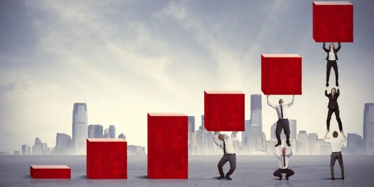 A conceptual digital illustration of professionals working together to build a rising bar chart out of large red blocks against a city skyline. The first three columns are solid red bars increasing in height. The fourth column shows a man holding up a block. The fifth column shows one man standing on another's shoulders to lift a block. The final, tallest column features a human tower of three people stacked vertically, lifting the highest red block.