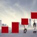A conceptual digital illustration of professionals working together to build a rising bar chart out of large red blocks against a city skyline. The first three columns are solid red bars increasing in height. The fourth column shows a man holding up a block. The fifth column shows one man standing on another's shoulders to lift a block. The final, tallest column features a human tower of three people stacked vertically, lifting the highest red block.